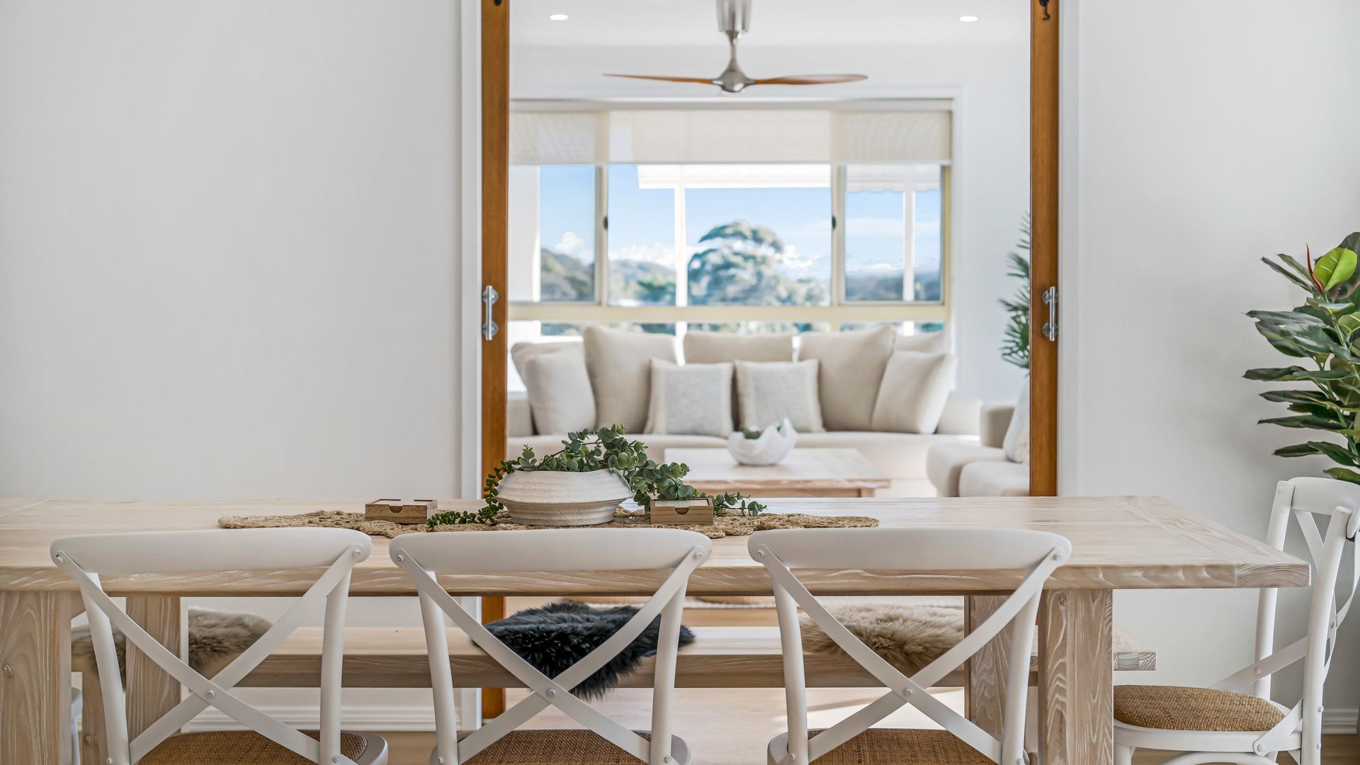 A dining area with a wooden table, white chairs, and a view of a living room with a sofa and a plant.