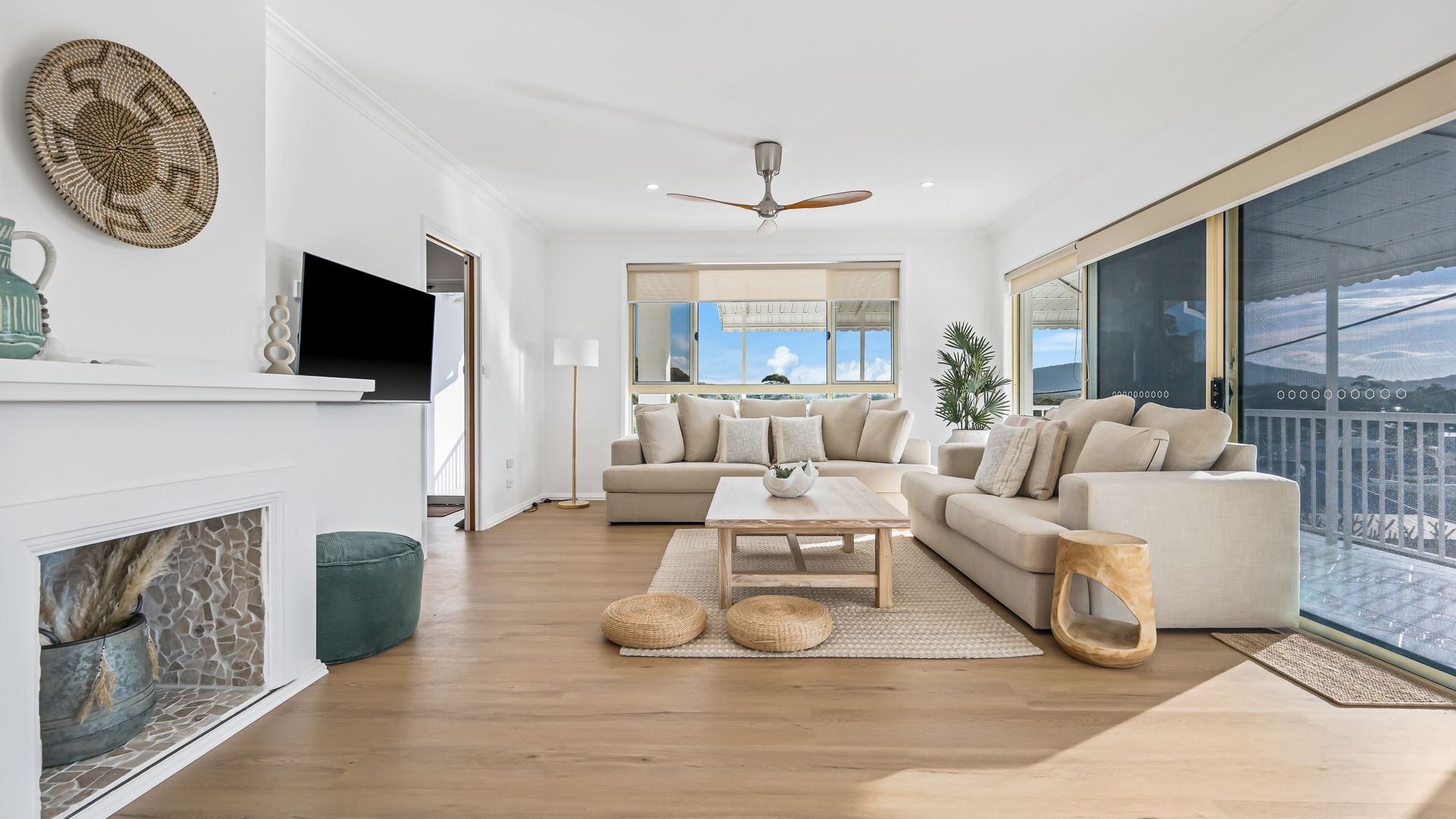 A modern living room with a fireplace, a large window with a view, a ceiling fan, and a sofa set.