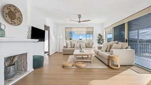 A modern living room with a fireplace, a large window with a view, a ceiling fan, and a sofa set.