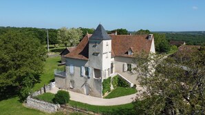 A large, historic stone house with a prominent tower, surrounded by lush greenery and a well-maintained garden.