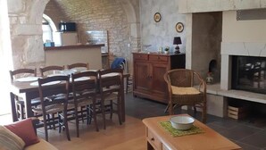 A cozy living room with a sofa, wooden coffee table, and a fireplace. There is a dining area with a table and chairs, and a kitchen area with a stone wall and a hanging light fixture.