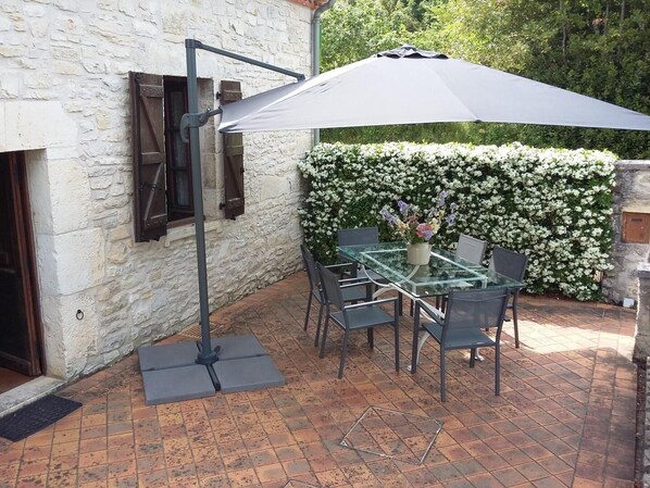 An outdoor patio with a glass table and chairs, a large umbrella, and a wall of white flowers.