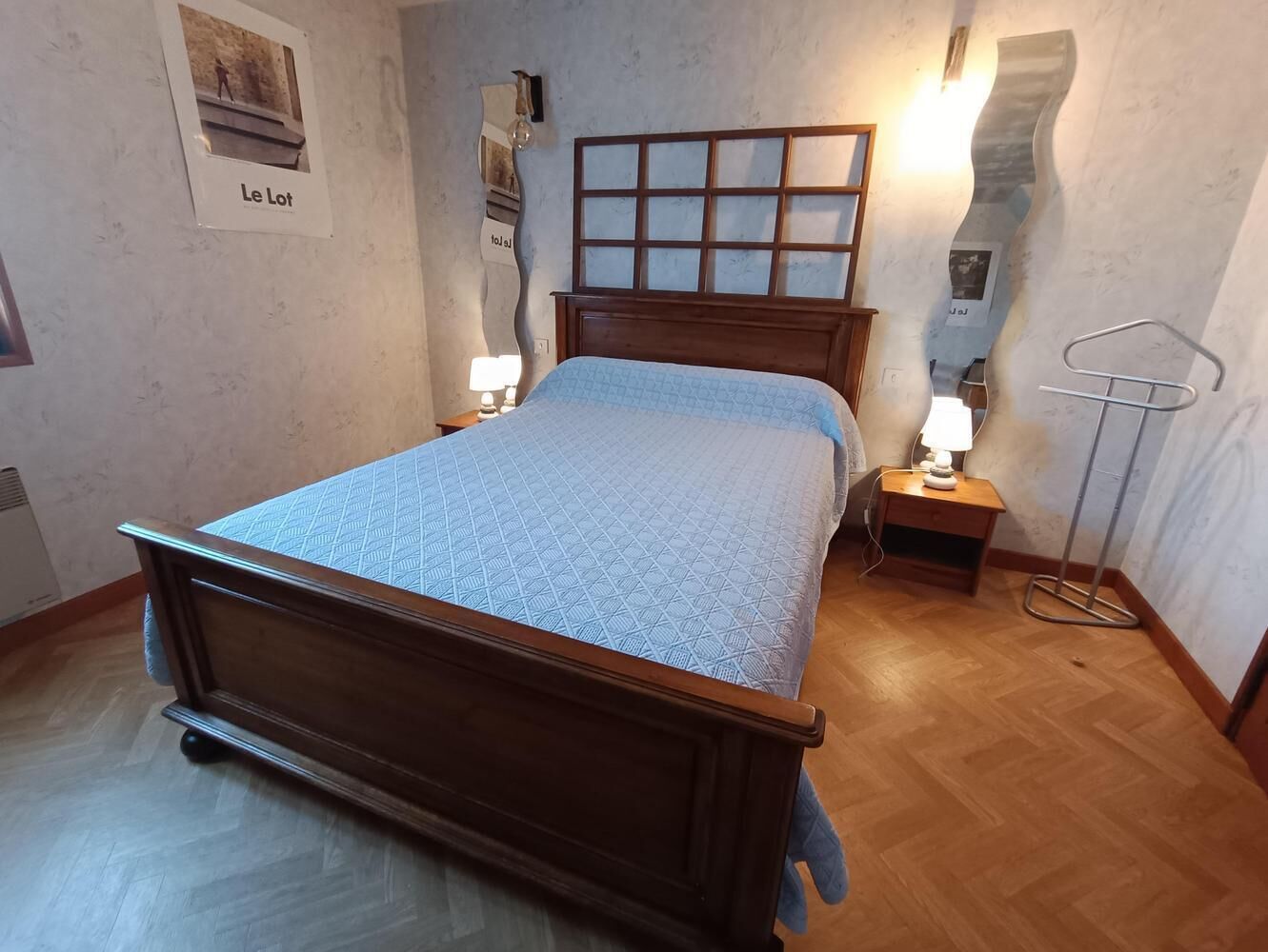 A bedroom with a wooden bed, a nightstand with a lamp, a wall-mounted mirror, and a framed picture labeled "Le Lot".