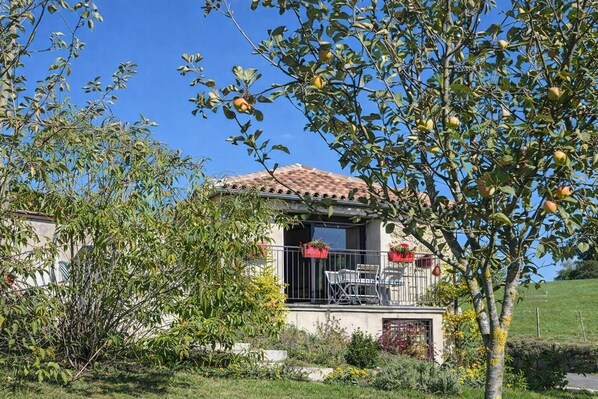 A house with a tiled roof, a balcony with chairs, and a garden with trees and shrubs.