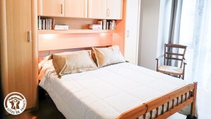 A bedroom with a bed, wooden headboard, built-in shelves with books, a chair, and a window with curtains. - Gîtes de France® - 46G12361 (VIAZAC)