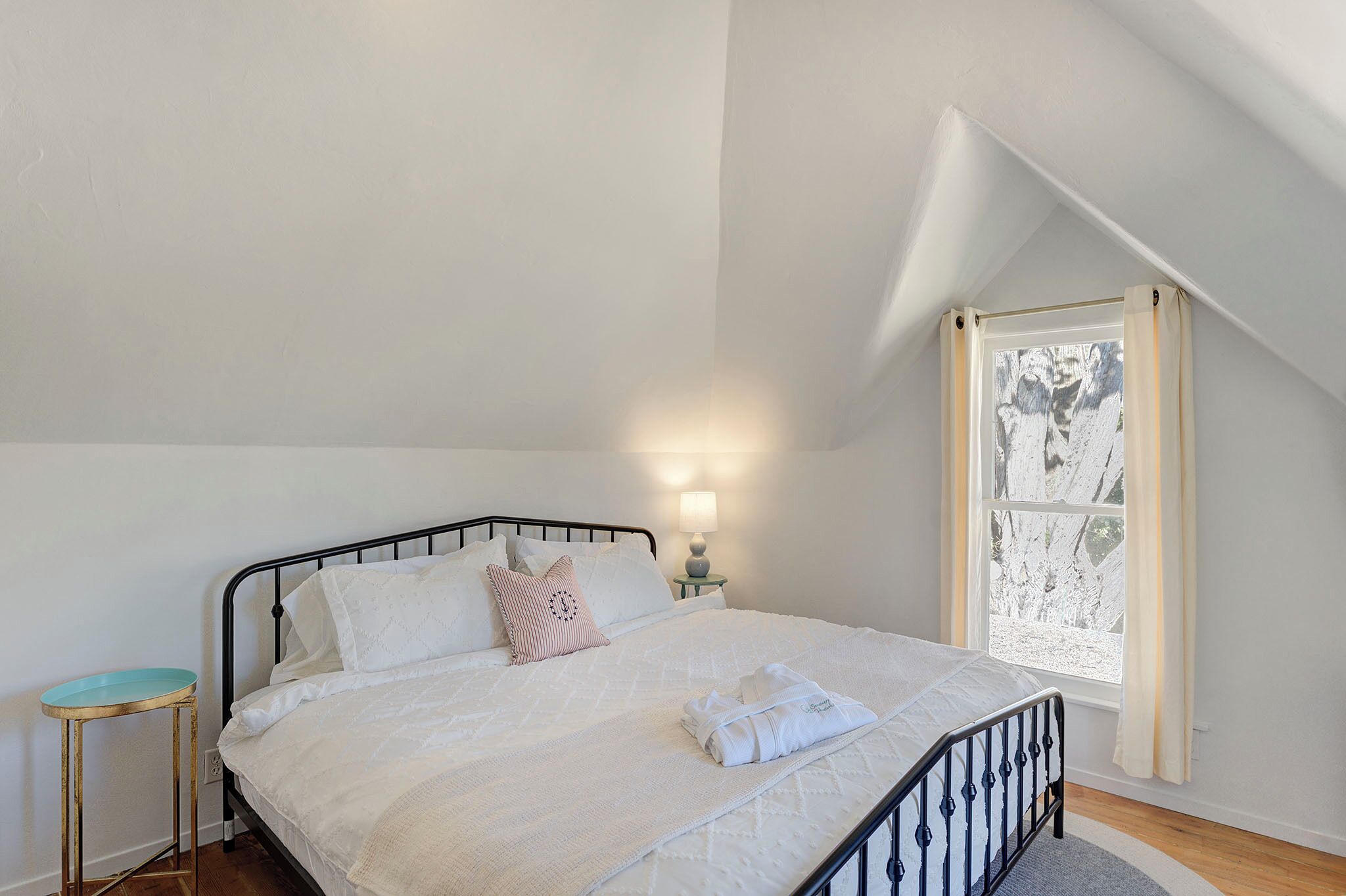 A neatly made bed with a white comforter and pillows, a bedside lamp, and a window with curtains.