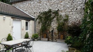 A stone building with a tiled roof, a white door, and a small window. There is a table with chairs and potted plants in the foreground. - Chez Yvette - Saint-sozy (SAINT-SOZY)