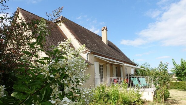 A two-story house with a sloped roof and a chimney, surrounded by lush greenery and flowering plants.