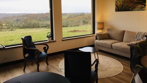 Ein großzügiges Wohnzimmer mit großen Fenstern und Blick auf eine grüne Landschaft. Es gibt ein Sofa, einen Sessel und einen kleinen Tisch.