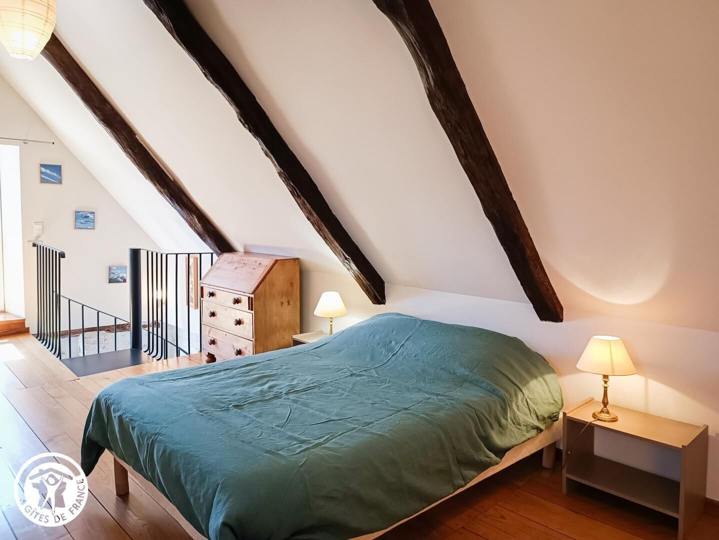 A bedroom with a sloped ceiling, wooden beams, a bed with a green bedspread, two bedside tables with lamps, and a wooden dresser.