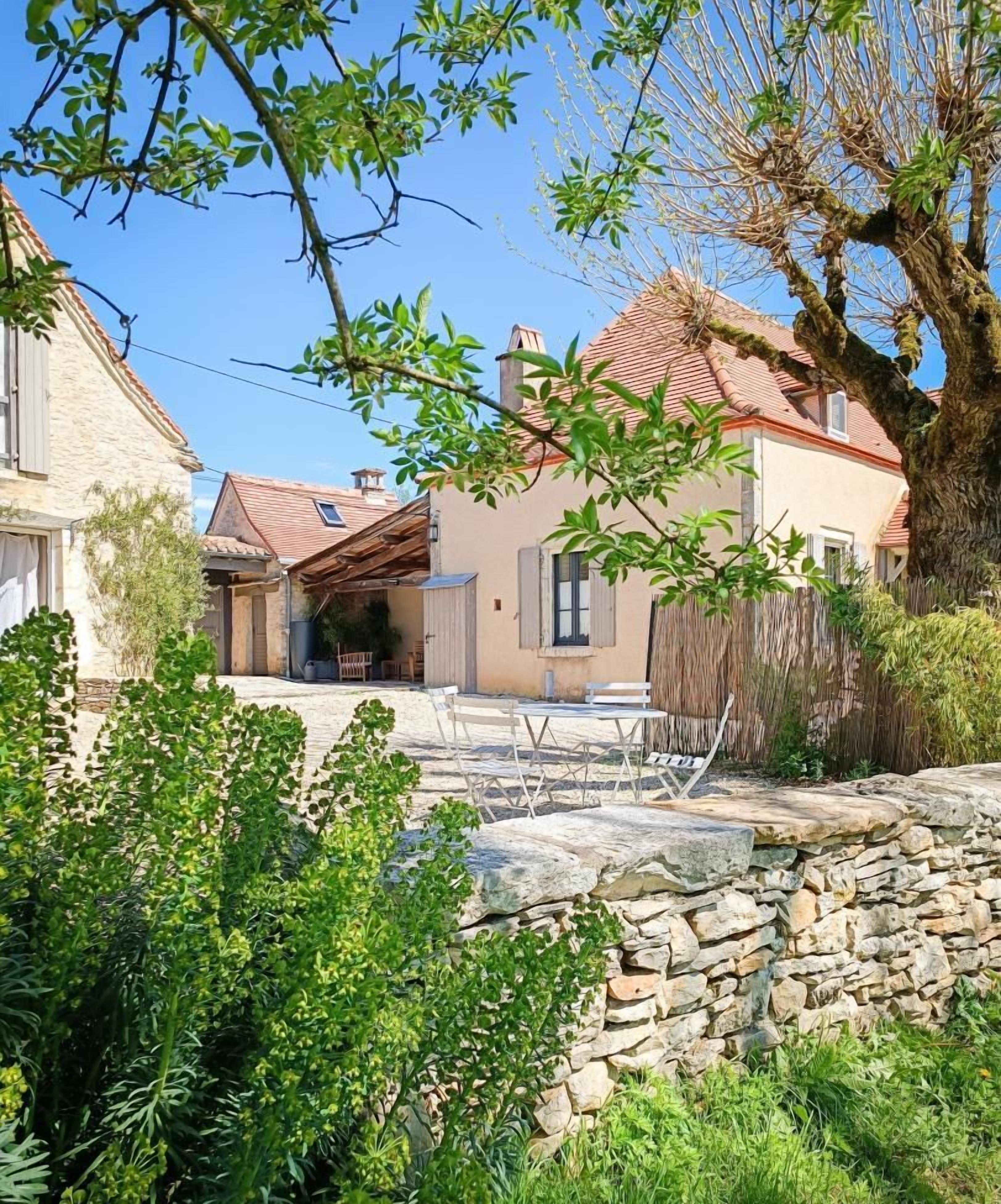 Un mur de pierre avec un espace patio, une clôture en bois et une maison avec un toit recouvert de tuiles rouges.