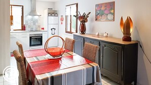 A modern kitchen with white cabinets, a built-in oven, and a refrigerator. There is a dining table with a red tablecloth and wicker chairs. A vase with flowers and a decorative light are on the counter.
