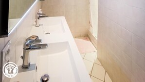 A bathroom with a large mirror, two sinks, a shower, and a small window.