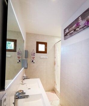 Un baño con un espejo grande, dos lavamanos, una ducha y un ventanal pequeño.