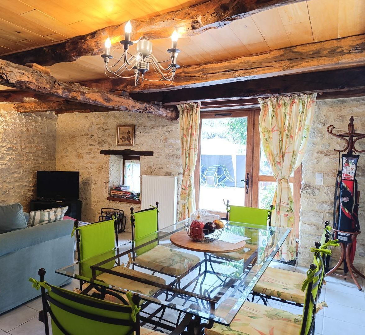A rustic room with a stone wall, wooden ceiling beams, a glass table with chairs, a sofa, a television, and a window with curtains.