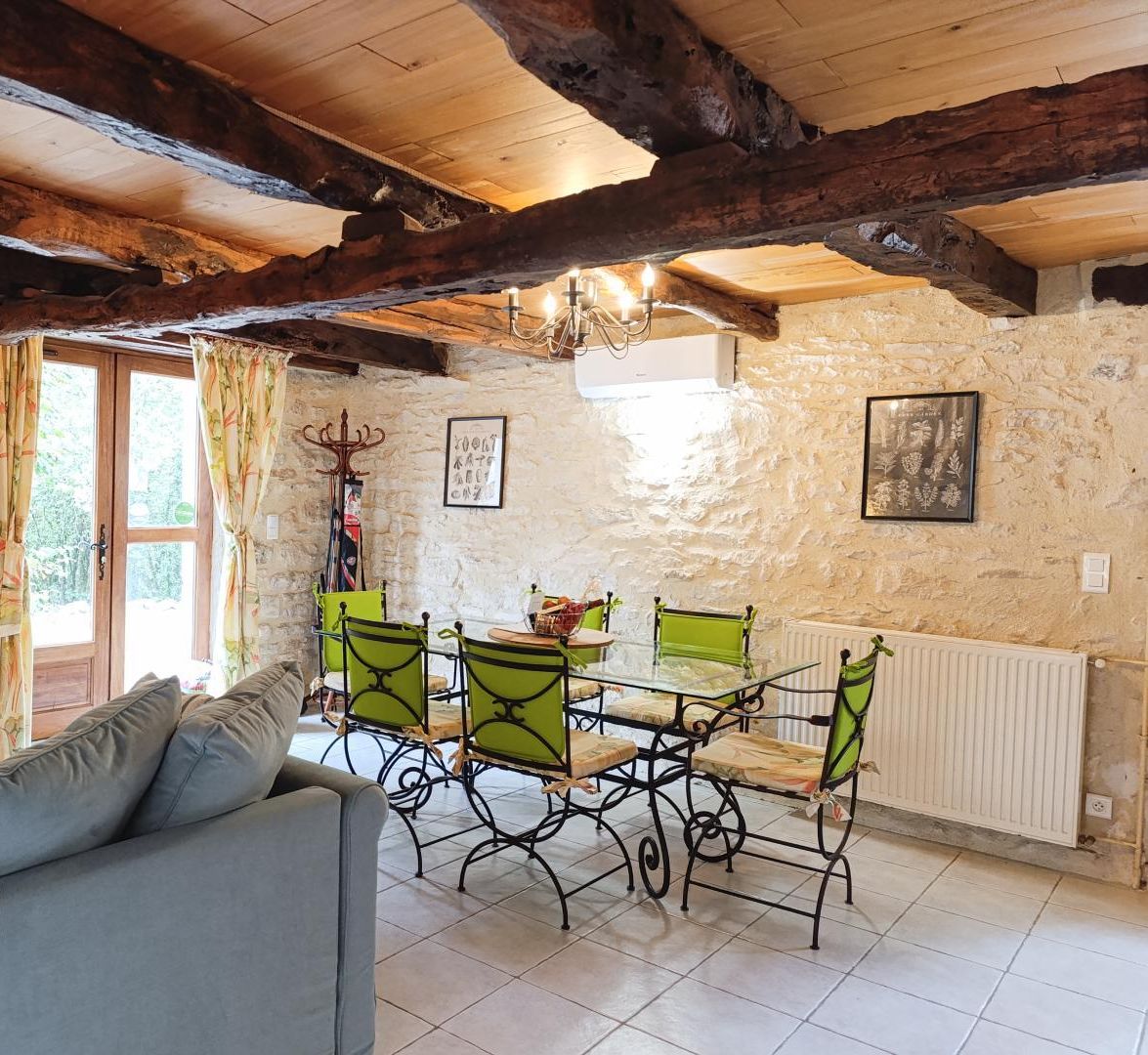 A rustic room with exposed wooden beams, stone walls, a dining table with green chairs, a sofa, and a chandelier.