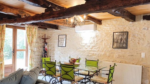 A rustic room with exposed wooden beams, stone walls, a dining table with green chairs, a sofa, and a chandelier.