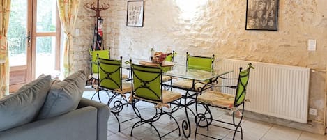 A rustic room with exposed wooden beams, stone walls, a dining table with green chairs, a sofa, and a chandelier.