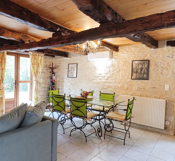 Une pièce rustique avec des poutres apparentes, des murs en pierre, une table à manger entourée de chaises vertes, un canapé et un lustre.