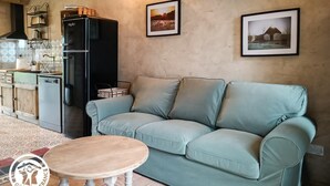En hyggelig stue med en lyseblå sofa, et lille træbord og et sort køleskab. To billeder hænger på væggen, og der er et køkken med vask og skabe.