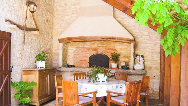 An outdoor patio with a stone wall, a brick oven, wooden furniture, and potted plants. - Gîtes de France® - Le Poutoy (SAINT-SIMON)