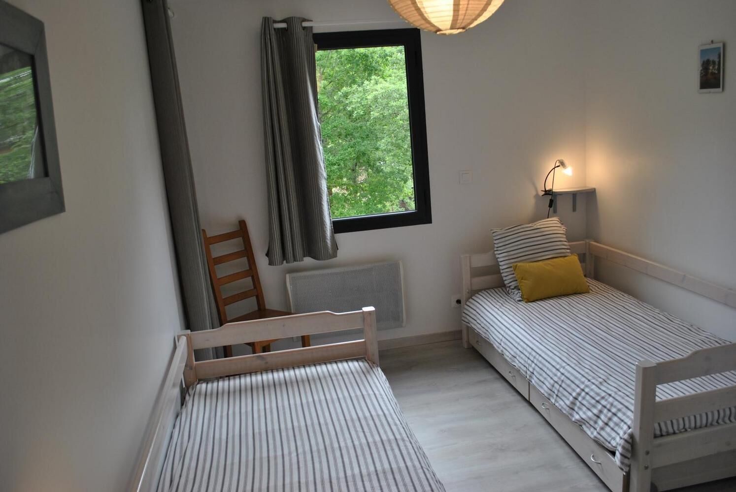 A bedroom with two single beds, a chair, a window with curtains, a radiator, and a wall-mounted lamp.