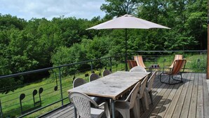 Een houten terras met een tafel en stoelen, omringd door een metalen hek, met uitzicht op een weelderig groen landschap.