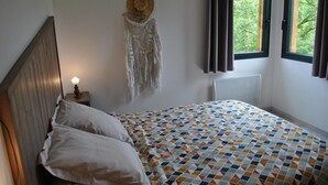 Een slaapkamer met een houten hoofdbord, een bed met een decoratieve spreidek, een wit kussen, een nachtkastje met een kaars, een macramé wandversiering en een raam met gordijnen.