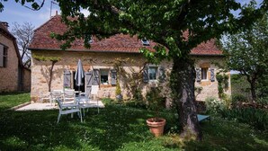 A stone house with a tiled roof, a patio with outdoor furniture, and a large tree in the foreground. - Gîtes de France® - LE MAS D'AYRENS (FIGEAC)