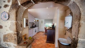 A kitchen with stone walls, a central island, and a view into the dining area. - Gîtes de France® - LE MAS D'AYRENS (FIGEAC)
