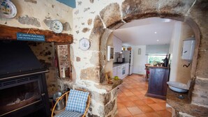 A rustic kitchen with a stone archway leading to a dining area, a charcoal grill, and a wicker chair. - Gîtes de France® - LE MAS D'AYRENS (FIGEAC)