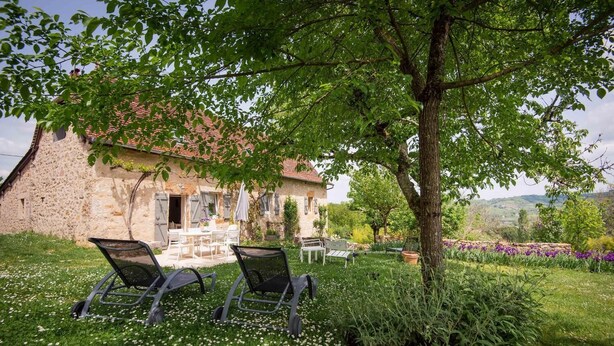 A stone building with a red-tiled roof, outdoor seating, and a garden with flowers and trees. - Gîtes de France® - LE MAS D'AYRENS (FIGEAC)