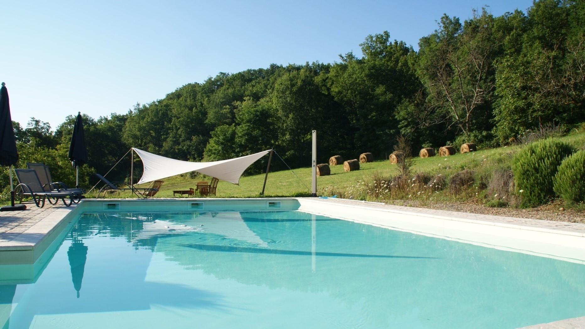 Una piscina con sillones de descanso y una pérgola, rodeada de césped y árboles.