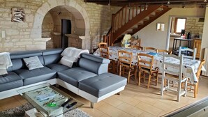 A cozy living room with a grey sectional sofa, a dining area with wooden chairs, and a stone wall with an archway. - Gîtes de France® - Gîte Le Cammas (SAINT SIMON)