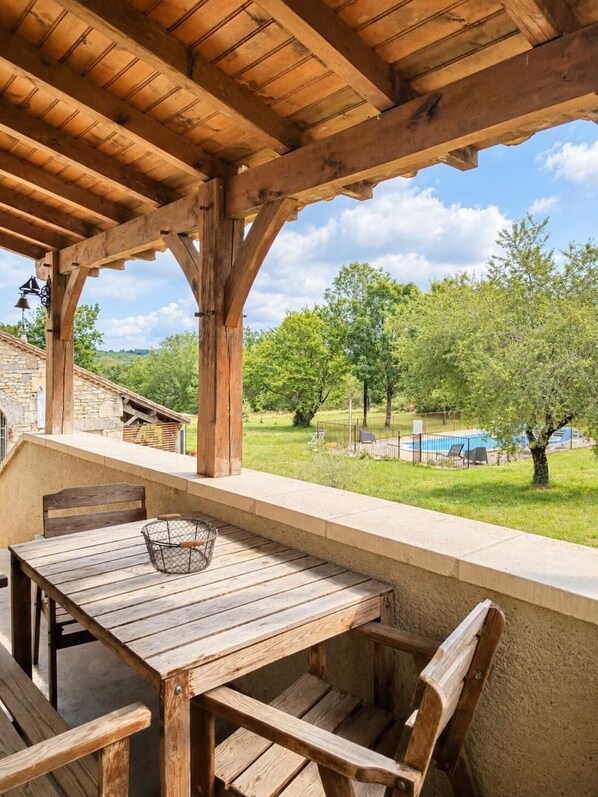 A wooden outdoor seating area with a table and chairs, overlooking a swimming pool and greenery.