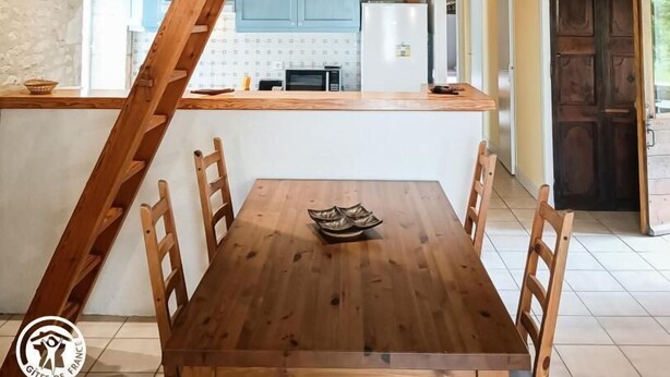 A rustic dining area with a wooden table and chairs, a kitchen in the background, and exposed wooden beams on the ceiling. - Coumbel - Villeseque (VILLESEQUE)