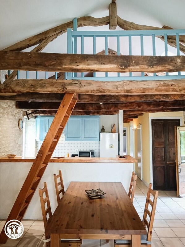 A rustic dining area with a wooden table and chairs, a kitchen in the background, and exposed wooden beams on the ceiling. - Coumbel - Villeseque (VILLESEQUE)