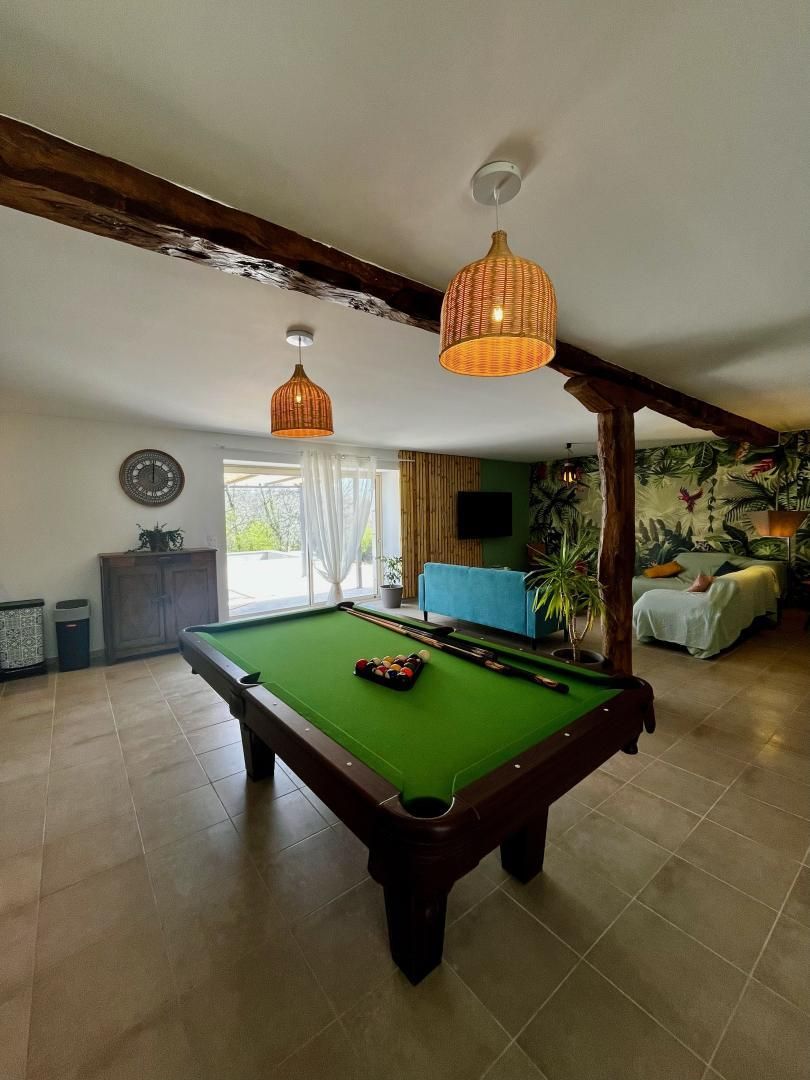 A pool table in a room with a wooden ceiling beam, a blue sofa, and a bed with a tropical wallpaper.