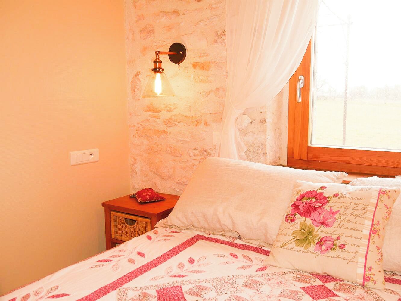 A bedroom with a bed, a nightstand, a wall-mounted lamp, and a window with curtains.