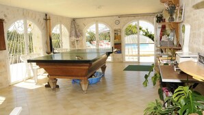Una mesa de billar en una habitación con amplios ventanales de arco, una piscina y un estante con diversos objetos.