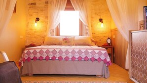 Un dormitorio acogedor con pared de piedra, una cama con colcha estampada, dos lámparas de pared y un ventanal con cortinas de encaje.