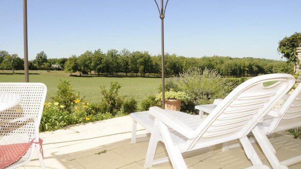 Un espacio de estar al aire libre cubierto, con sillones blancos y una mesa, con vistas a un campo verde y árboles.