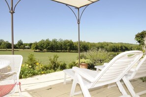 A covered outdoor seating area with white lounge chairs and a table, overlooking a green field and trees.