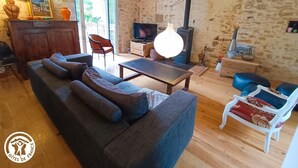 A cozy living room with a sofa, chairs, a TV, a fireplace, and a wooden ceiling. - Gîtes de France® - Le Fournil (GIGOUZAC)