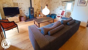 A cozy living room with a stone wall, a wooden floor, a sofa, a TV, a fireplace, and a hanging light fixture. - Gîtes de France® - Le Fournil (GIGOUZAC)