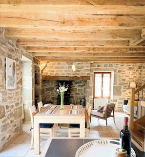 A rustic room with a stone fireplace, wooden beams, a dining table with chairs, and a sofa.