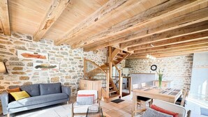 Un interior rústico con vigas de madera a la vista, una pared de piedra, un sofá, una mesa de comedor y una escalera.