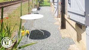 A narrow pathway leading to a building with a large wooden door, a small table with a chair, and a garden area with plants and flowers.