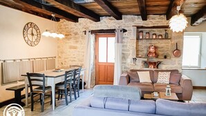 A rustic interior with exposed wooden beams, a stone wall, a dining area with a table and chairs, and a seating area with a sofa and cushions. - Gîtes de France® - La Caze (ALVIGNAC LES EAUX)