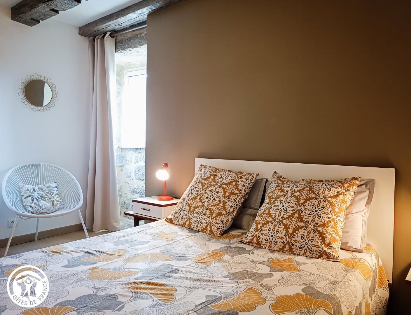 Um quarto com uma cama, duas almofadas estampadas, uma mesa de cabeceira com abajur, uma cadeira branca e um espelho redondo na parede.
